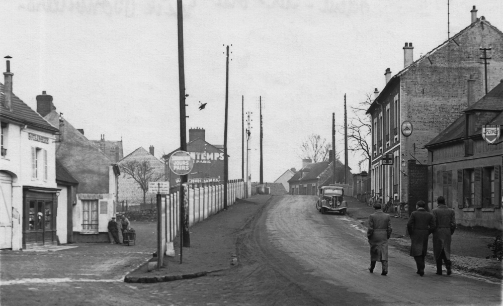 Nogent-sur-Oise. La rue de Bonvillers, dans la montée du pont passant au-dessus de la voie ferrée. Le 57 bis est peut-être dans un groupe de maisons situé derrière la pharmacie, accessible par un passage perceptible au-delà des trois hommes. Carte postale. Collection Mémoire Vive.