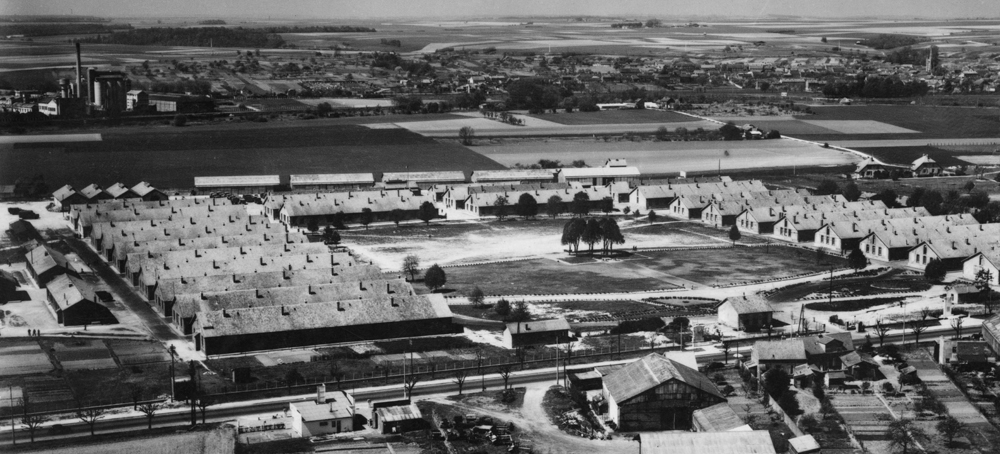 La caserne de Royallieu après-guerre. Les huit premiers bâtiments alignés à gauche sont ceux du quartier “A”, désigné pendant un temps comme le “camp des communistes”. À l’arrière plan à gauche, sur l’autre rive de l’Oise, l’usine de Venette qui fut la cible de plusieurs bombardements avec “dégâts collatéraux” sur le camp. Carte postale. Collection Mémoire Vive.