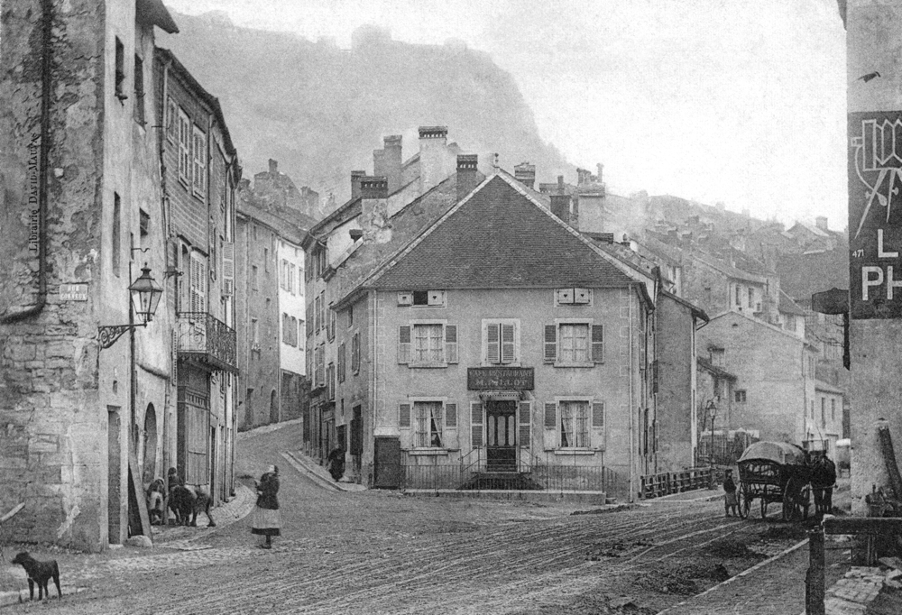 Salins-les-Bains. Le café-restaurant à l’angle de la rue d’Olivet, à gauche, et de l’avenue Gambetta, à droite. En surplomb à l’arrière plan, le fort Belin. Carte postale des années 1900, collection Mémoire Vive.